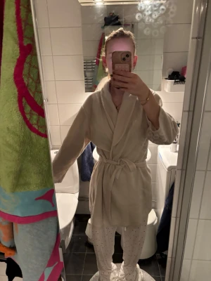 Beige fleecemorgonrock med knytband - Mysig beige morgonrock i fleece med omlottkrage och knytband i midjan. Perfekt för att ha hemma. Enkel och stilren design utan mönster eller extra detaljer.