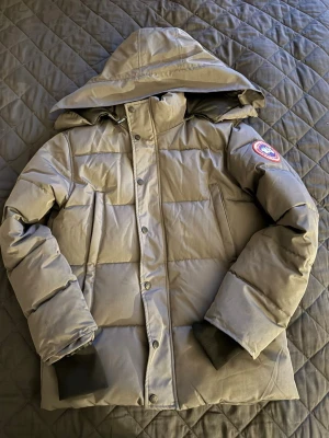 Grå dunjacka från Canada Goose - Grå dunjacka från Canada Goose med huva och klassisk logga på ärmen. Jackan har lång ärm, knappar och dragkedja framtill. Perfekt för kalla dagar. 