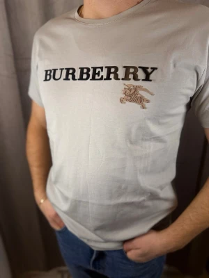 Grå Burberry t-shirt med logga - Snygg vit t-shirt från Burberry med stor svart logga och klassiskt riddarmotiv på bröstet. T-shirten har rund halsringning och korta ärmar. Tillverkad i mjuk bomull som känns skön mot huden. Perfekt för dig som vill ha en clean och stilren look.