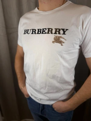 Vit Burberry t-shirt med logga - Snygg vit t-shirt från Burberry med stor svart logga och klassiskt ridmotiv på bröstet. T-shirten har rund halsringning och är kortärmad. Tillverkad i mjuk bomull som känns skön mot huden. Perfekt för dig som vill ha en clean och stilren look.