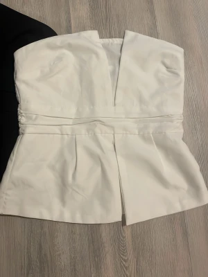 Svart och vit axelbandslös korsettopp - Säljer två stilrena axelbandslösa korsettoppar, en i svart och en i vit. Båda har markerad midja, snygga veckdetaljer och en djup v-ringning framtill. Topparna stängs med dragkedja bak och har en lätt utsvängd nederdel. Perfekta att styla till jeans eller kjol.