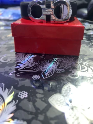 Svart skärp från Salvatore Ferragamo - Säljer ett svart skärp från Salvatore Ferragamo med ikoniskt silverfärgat spänne. Skärpet är tillverkat i äkta skinn och har en stilren, klassisk design. Kommer i original röd låda med guldfärgad logga. Perfekt accessoar för att lyfta din outfit.