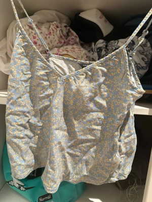 Blommig blå linnetopp med smala band - Supersöt blå linnetopp från Lindex med ljusgult blommönster. Toppen har tunna justerbara axelband och v-ringad hals. Perfekt till sommaren och ger en fräsch vibe till din outfit.