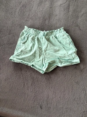 Ljusgröna shorts från H&M med snörning - Säljer ett par ljusgröna shorts från H&M i mjuk bomull. Shortsen har elastisk midja med snörning och söta volangdetaljer vid fickorna. Perfekta för varma dagar och riktigt sköna att ha på sig.