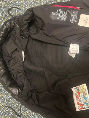 Svart Moncler cardigan - Säljer en svart pufferjacka från Moncler med glansig, quiltad framsida och stickade ärmar. Jackan har huva med röd och vit detalj, dragkedja framtill och Moncler-logga på axeln. Perfekt för kyliga dagar och riktigt snygg streetstil.