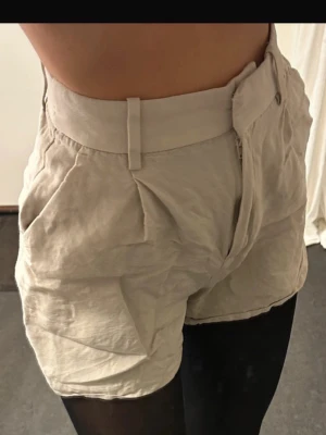 Beige högmidjade shorts med fickor - Säljer ett par beige shorts med hög midja och bred linning. Shortsen har snygga veck framtill och praktiska fickor på sidorna. De är tillverkade i ett lätt och mjukt tyg, perfekta för varmare dagar. Stängs med dragkedja i sidan och har hällor för bälte.