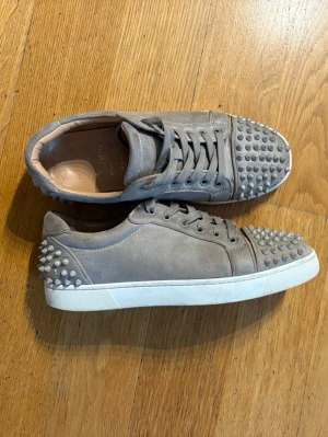 Beiga mocka sneakers med nitar - Säljer ett par beiga sneakers från Christian Louboutin i mocka med coola nitar på tå och häl. Skorna har snörning och den klassiska röda sulan som sticker ut. Perfekta för dig som vill ha något unikt och edgy i garderoben.