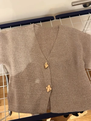 Beige ribbad kofta med kort ärm - Kortärmad beige kofta med ribbad struktur och två unika träknappar framtill. V-ringad och i mjukt stickat material, perfekt för lager på lager under vår och höst. Enkel och stilren design som passar till det mesta.