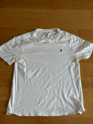 Vit t-shirt från Polo Ralph Lauren XL - Klassisk vit t-shirt från Polo Ralph Lauren i storlek XL. T-shirten har korta ärmar, rund hals och den ikoniska broderade loggan på bröstet. Tillverkad i mjuk bomull som känns skön mot huden. Perfekt basic-plagg med clean look.