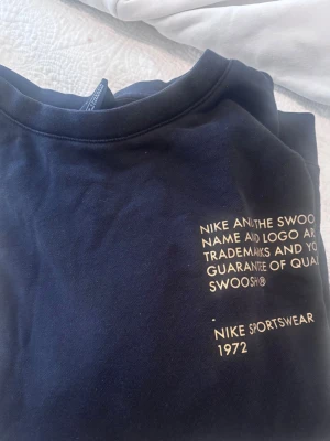 Svart Nike sweatshirt med guldtryck - Nike sweatshirt i svart med stora guldfärgade SWOOSH-tryck på ärmen och bröstet. Klassisk rund hals och långa ärmar. Tillverkad i mjuk bomullsmix, perfekt för en sportig och chill look. Märkt med Nike Sportswear 1972 och ikoniska swoosh-loggor.