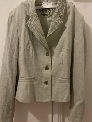 Beige kavaj från AD Fashion - Snygg beige kavaj från AD Fashion med markerad krage och dekorativa sömmar. Kavajen har tre knappar framtill, raka fickor och är tillverkad i ett lätt linneliknande material. Perfekt för dig som vill ha en stilren och klassisk look.