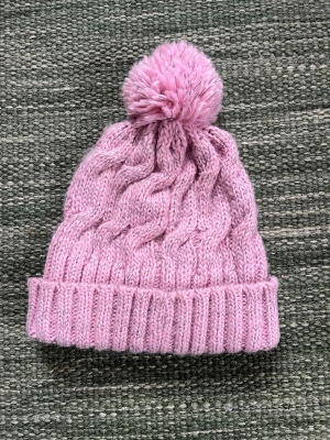 Rosa stickad mössa med tofs Lindberg - Mysig rosa stickad mössa med reflexgarn från Lindberg med stor tofs på toppen och bred vikt kant.  Insidan är fodrad med mjuk fleece för extra värme. Perfekt för kalla dagar och ger en söt look till vinteroutfiten.