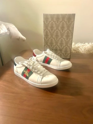 Gucci Ace vita sneakers med bi-detalj - Gucci Ace sneakers i vitt skinn med klassiska gröna och röda ränder på sidan samt broderad guldfärgad bi. Baksidan har metallicröd detalj på ena skon och metallicgrön på den andra, båda med Gucci-logga. Platt sula och snörning framtill.