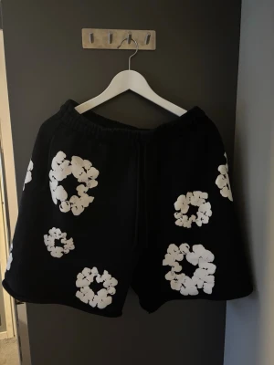 Svarta shorts Denim Tears blommig M - Svarta shorts från Denim Tears i storlek M med vita blommönster över hela plagget. De har elastisk midja med snörning och är tillverkade i mjuk bomull. Perfekta för en chill och trendig look.