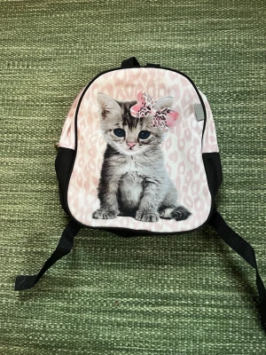 Rosa barnryggsäck med kattmotiv från Mywear - Supersöt ryggsäck i rosa och svart från Mywear med ett stort tryck av en kattunge med rosett framtill. Väskan har justerbara axelband och dragkedja. Perfekt för dig som vill ha en gullig och unik accessoar till förskolan eller stan.