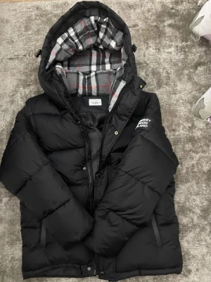 Svart pufferjacka från Burberry - Säljer en svart pufferjacka från Burberry med avtagbara ärmar så den kan användas som väst. Jackan har huva med rutigt foder i grått, vitt och rött samt tydlig Burberry-patch på ärmen. Stängs med dragkedja och knappar. Perfekt för kalla dagar.