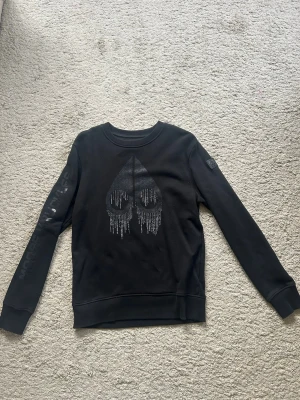 Svart sweatshirt Moose Knuckles - Säljer en svart sweatshirt från Moose Knuckles med ett silverfärgat tryck framtill och logga på ärmen. Tröjan har rund hals, långa ärmar och är i storlek Small. Tröjan är använd men fortsatt i bra skick, köpt på Zgander i Helsingborg.