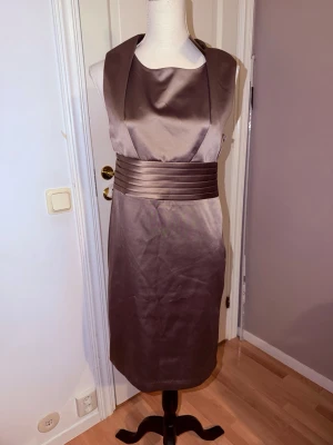 Brun satin klänning från Stockh LM - Supersnygg entire brun klänning från Stockh LM i satin med bred markerad midja och hög krage. Klänningen är ärmlös och har en elegant sopad siluett som ger en lyxig känsla. Perfekt för dig som vill sticka ut med en stilren look.