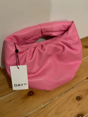 Rosa axelväska från DAY ET - Säljer en superfin rosa axelväska från DAY ET i mjukt syntetmaterial. Väskan har en cool puffig form, dragkedja upptill och ett stilrent DAY ET-logo i silver på framsidan. 