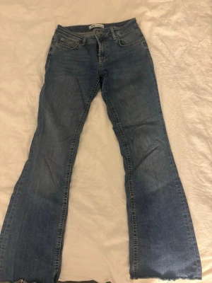 Blå bootcut jeans från Zara - Säljer ett par klassiska blå jeans från Zara i bootcut-modell. Jeansen har normal midja, fem fickor och råa kanter längst ner på benen för en avslappnad vibe. Tillverkade i mjukt bomullsjeans-tyg och perfekta för dig som gillar en tidlös look.