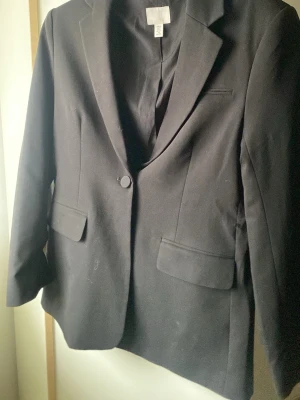 Svart kavaj från H&M XS - Klassisk svart kavaj från H&M i storlek XS. Enkelknäppt modell med två fickor fram och snygg krage. Perfekt för en clean och stilren look. Materialet känns mjukt och följsamt, och passformen är normal.