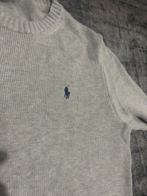 Ljusgrå stickad tröja Polo Ralph Lauren - Snygg ljusgrå stickad tröja från Polo Ralph Lauren med klassisk blå logga broderad på bröstet. Tröjan har rund halsringning, ribbade muddar och är långärmad. Perfekt för dig som vill ha en clean och stilren look. Köptes också is USA men aldrig använt