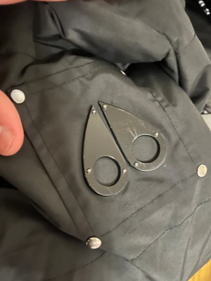 Svart dunjacka från Moose Knuckles - Säljer en svart dunjacka från Moose Knuckles med ikoniska logotypen i metall på ärmen och coolt broderat foder med hockeymotiv. Jackan har huva, dragkedja och flera fickor med snygga detaljer. Perfekt för kalla dagar och riktigt street.