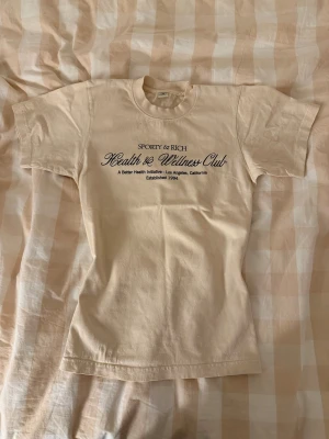 Beige t-shirt Sporty & Rich XS - Beige t-shirt från Sporty & Rich med svart textprint på bröstet. Klassisk rund halsringning och korta ärmar. Tillverkad i mjuk bomull och har en clean, stilren look. Perfekt för dig som gillar minimalistisk och sportig stil. Använd fåtal gånger. 