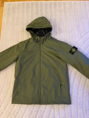 Grön vindjacka från Stone Island - Säljer en grön vindjacka från Stone Island med ikonisk patch på ärmen. Jackan har huva med snörning, dragkedja framtill och två sidofickor. Materialet är lätt och vattenavvisande, perfekt för blåsiga dagar. Snygg och funktionell design som sticker ut.