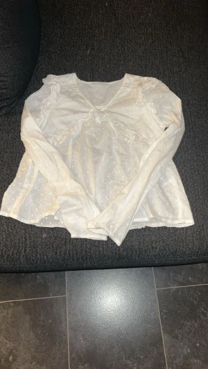 Vit prickig blus med volangdetaljer - Säljer en söt vit blus med diskreta prickar och volangdetaljer vid axlarna. Blusen har v-ringad hals och långa ärmar, perfekt för dig som gillar romantisk och feminin stil. Materialet känns lätt och luftigt, vilket gör den skön att bära.