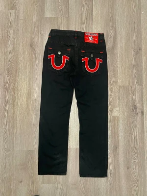 True Religion svarta jeans med röda detaljer  - Svarta jeans från True Religion, modell Joey Super T. Använde dom 2-3 gånger. Snygga kontrastsömmar i rött och silverfärgade knappar. Bakfickorna har ikonisk broderad logga i rött och vitt. Klassisk rak passform och robust jeansmaterial. Perfekt för dig som gillar streetwear med attityd.   Längd: 112 cm Midja: 42 Raise: 27 Lår: 26