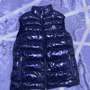 Svart glansig dunväst från Moncler - Snygg svart dunväst från Moncler med glansig finish och hög krage. Västen har två dragkedjefickor framtill och stängs med dragkedja. Klassisk Moncler-logga på bröstet. Perfekt för lager-på-lager och streetstyle.