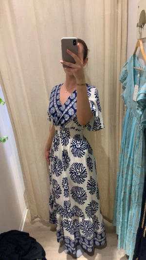 Vit och marinblå långklänning med mönster - Säljer en superfin långklänning i vit och marinblå med blommigt mönster. Klänningen har v-ringad omlott upptill, korta ärmar och ett smockat midjeparti som ger en snygg siluett. Materialet känns som bomull, perfekt för varma dagar.