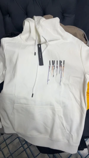 Amiri Hoodie  - Säljer en vit hoodie från Amiri med coolt färgstänksmönster och stor logga både fram och bak. Tröjan har huva med snörning och en stor magficka. Materialet är mjuk bomull och passformen är normal. Perfekt för dig som gillar streetwear och vill sticka ut. Och storlek är M