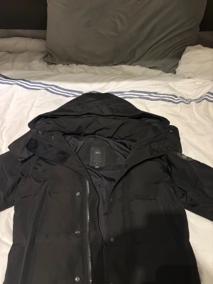 Svart pufferjacka från Canada Goose - Säljer en svart pufferjacka från Canada Goose med huva och snyggt märke på ärmen. Jackan har dragkedja och knappar framtill, samt flera praktiska fickor. Perfekt för kalla dagar och har en stilren look som funkar till allt.