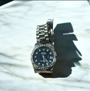  silver klocka -  klocka i silver med rund boett och länkarmband i metall. Urtavlan är mörk med  markerade index och datumvisning. Ringen runt boetten har dekorativa detaljer och ej äkta  baksidan lite sönder  visar den automatiska mekanismen och rost genom ett transparent fönster. Klassisk och lyxig design.