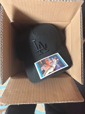 Svart LA-keps från New Era 9FORTY - Snygg svart keps från New Era 9FORTY med LA-logga broderad framtill. Skärmen är röd på undersidan och kepsen har justerbar passform. Perfekt för dig som gillar sportig stil och baseball. Materialet är bomull och polyester.