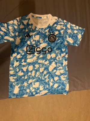 Ajax blå och vit fotbollströja Adidas S - Ajax fotbollströja i storlek S från Adidas med ett unikt blått och vitt mönster. Tröjan har korta ärmar, rund hals och Ajax-emblem med tre stjärnor på bröstet. Materialet är lätt polyester och Ziggo-loggan syns tydligt framtill.