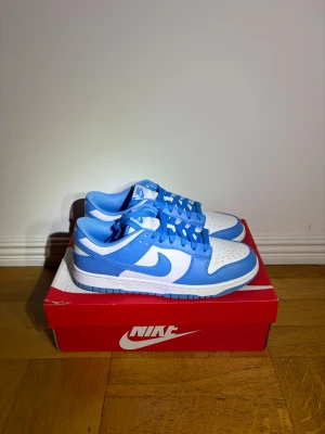 Nike Dunk Low blå och vita sneakers - Helt nya och oanvända Nike Dunk Low sneakers i fräsch blå och vit färg. Skorna har klassisk låg siluett, rund tå och snörning. Ovandelen är i skinn med perforering på tån för extra ventilation. Snygg kontrast mellan det blå och vita, med Nike-logga på sidan och tungan.