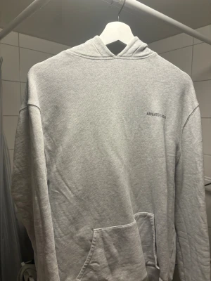 Ljusgrå hoodie från Arigato - Säljer en stilren ljusgrå hoodie från Arigato med diskret logga på bröstet. Tröjan har huva, lång ärm och en klassisk magficka framtill. Perfekt för dig som gillar enkel och clean streetwear.