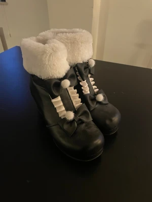 Svarta boots med vit fuskpäls och rosetter - Svarta lolita boots med vit fuskpäls runt skaftet, dekorerade med svarta rosetter. Skorna har klack och dragkedja på sidan, samt vita veckade detaljer framtill. Perfekt för dig som gillar japanskt mode och söta detaljer.