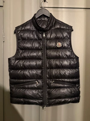 Svart dunväst från Moncler - Svart dunväst från Moncler med quiltad design och hög krage. Västen har två dragkedjeförsedda fickor framtill och Moncler-logga på bröstet. Materialet är glansigt och lätt, perfekt för lager-på-lager. Stängs med dragkedja hela vägen.