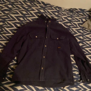 Mörkblå overshirt  - Säljer en mörkblå overshirt med klassisk krage och två stora bröstfickor med knappar. Skjortan har långa ärmar med manschett och knappar framtill. Perfekt för dig som gillar en enkel och stilren look. Den är storlek XXS. Etikett finns ej.