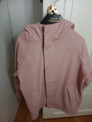 Ljusrosa regnjacka med huva - Säljer en ljusrosa regnjacka med huva och praktiska fickor framtill. Jackan har knappar längs hela framsidan och är tillverkad i ett slätt syntetmaterial som står emot regn. Perfekt för dig som vill ha en clean och chill look när det är blött ute.