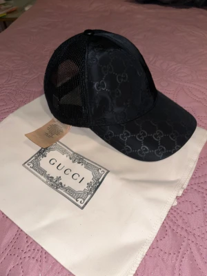 Svart Gucci keps med mesh och logga - Snygg svart keps från Gucci med klassisk GG-monogram över hela framsidan och mesh på sidorna och bak. Justerbar snapback och läderdetalj med Gucci-logga baktill. Tillverkad i Italien, stilren och lyxig design som sticker ut.