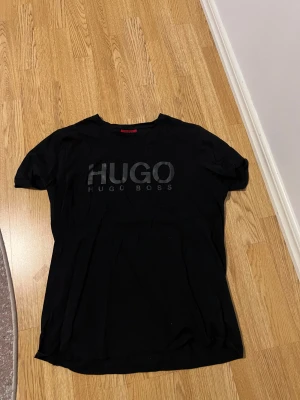 Svart t-shirt från Hugo Boss - Svart t-shirt från Hugo Boss med stor HUGO-logga i svart på bröstet. Klassisk rund hals och korta ärmar. Tillverkad i mjuk bomull som känns skön mot huden. Perfekt för dig som gillar stilrena och enkla plagg med tydlig branding.