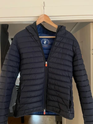 Mörkblå pufferjacka Save The Duck - Säljer en mörkblå pufferjacka från Save The Duck med huva och dragkedja. Jackan är quiltad och tillverkad i syntetmaterial, helt animal free. Perfekt för dig som vill ha en lätt och stilren jacka med schysst passform. Tål kyla väldigt bra. Nypris 1700 kr