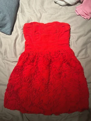 Röd axelbandslös kortklänning - Säljer en röd axelbandslös kortklänning med volangdetaljer och rosformade texturer på kjolen. Klänningen har en figurnära passform upptill och en fluffig kjol som ger en festlig look. Perfekt för bal eller student. Materialet känns mjukt och har struktur.