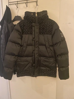 Svart pufferjacka Glorious Gangsta XS - Svart pufferjacka från Glorious Gangsta med fet geometriskt mönster i sammet på axlar, huva och nederkant. Jackan har fluffig fuskpäls på huvan, dragkedja framtill och flera fickor. Perfekt för kalla dagar med sin tjocka vaddering och coola detaljer. Jackan är i storlek XS men passar även S, använd ett par fåtal gånger. Pris kan diskuteras 