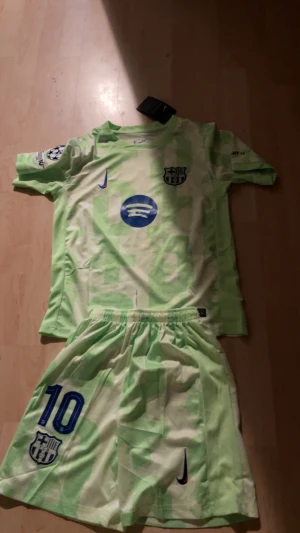 Barcelona fotbollströja och shorts - Säljer ett Barcelona fotbollskit med tröja och shorts i ljusgrön färg med mönster. Tröjan har korta ärmar, klubbmärke, Nike-logga och Spotify som sponsor. Shorts med nummer 10 och klubbmärke. Perfekt för dig som älskar Barca och vill sticka ut på planen.  Lamine Yamal nummer 10